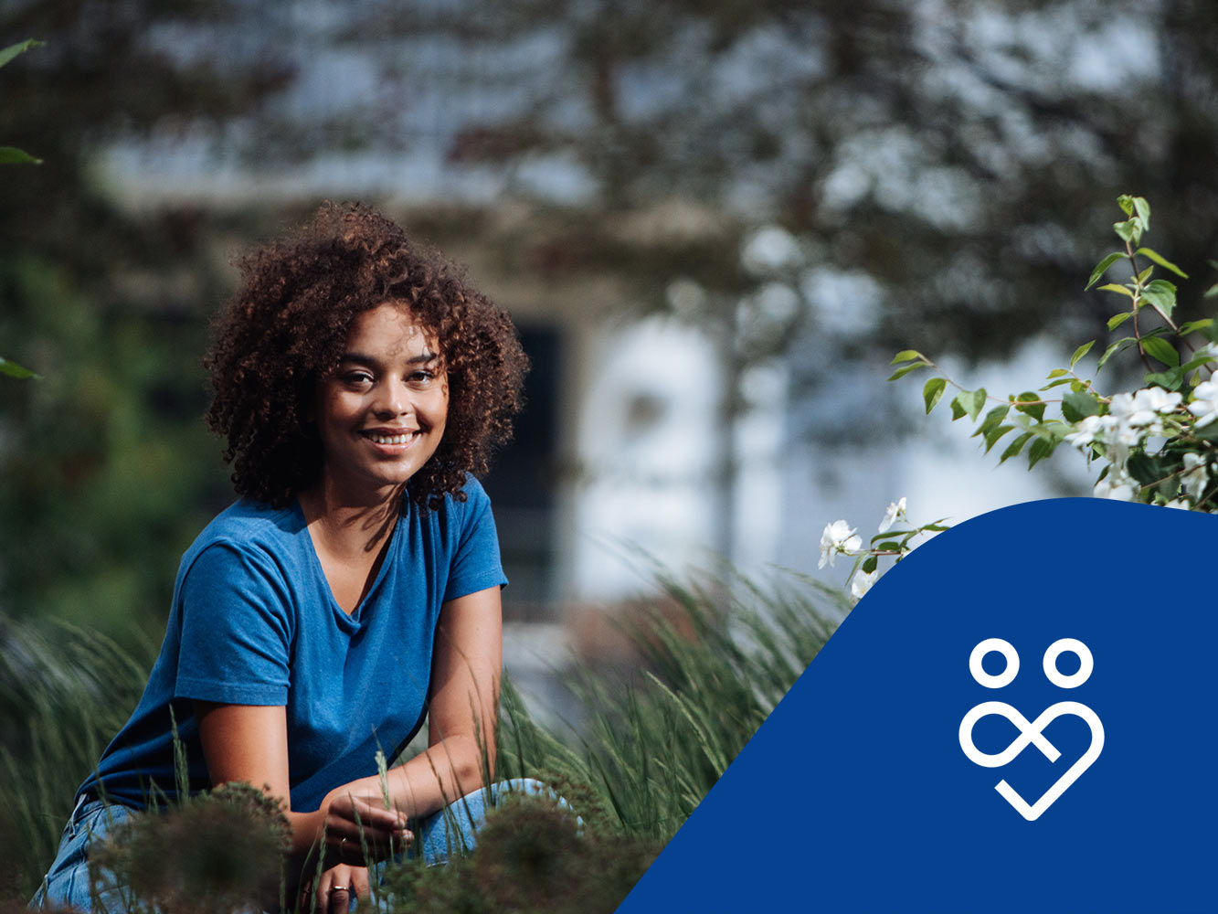 Kvinna sitter på huk i gräset med en fastighet i bakgrunden. Trygghetsikon.