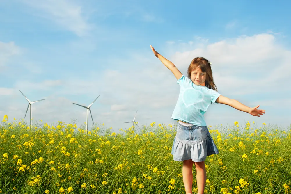 Flicka står med utsträckta armar i ett rapsfält med vindkraftverk bakom.